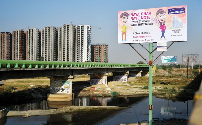 Bảng quảng cáo của một nhà môi giới bất động sản với hình ảnh một nhân vật cầm tờ tiền 2.000 rupee mới cùng slogan “Nhà mới, tiền mới”, gần khu vực đang xây dựng các tòa chung cư tại Uttar Pradesh
